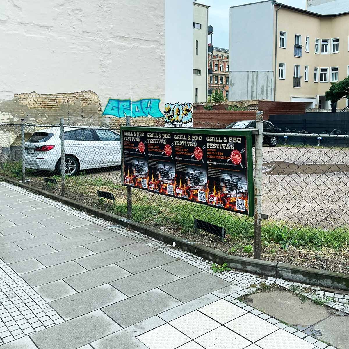 Plakatwand mit Ankündigungen für ein Grill- und BBQ-Festival, umgeben von einem Zaun.