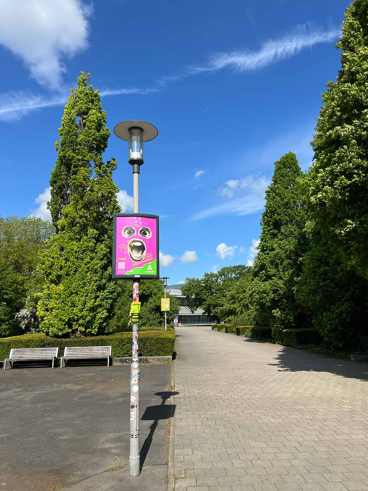 Öffentliche Informationssäule mit einem bunten Plakat in einem Park, umgeben von Bäumen und einem klaren Himmel.