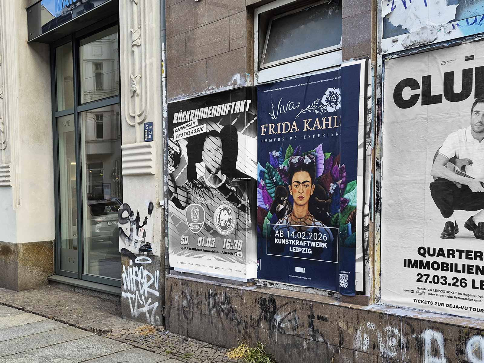 Straßenwand mit bunten Plakaten, darunter Viva Frida Kahlo Immersive Experience am Kunstkraftwerk Leipzig; weitere Poster und Graffiti an einer Fassadenwand