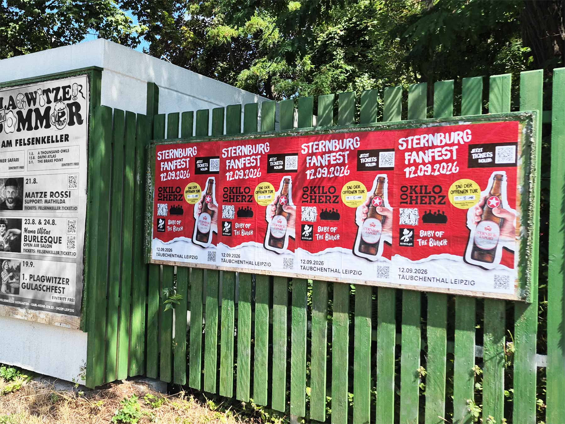 Mehrere rote Sternburg-Fanfest-Plakate hängen an einem grünen Holzzaun vor Bäumen in Leipzig.