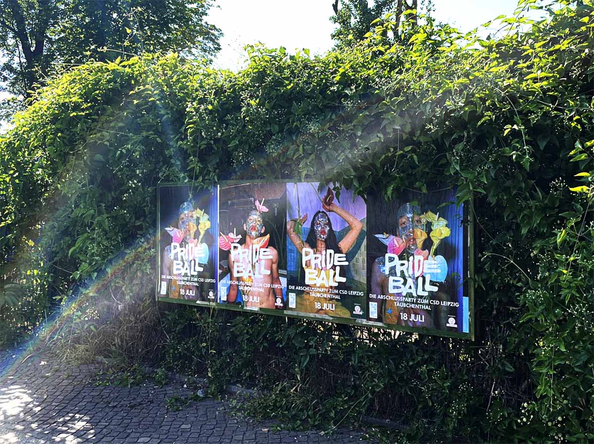 Entlang einer begrünten Wand hängen vier bunte Prideball-Plakate mit der Aufschrift „Pride Ball“ und dem Datum 18. Juli.